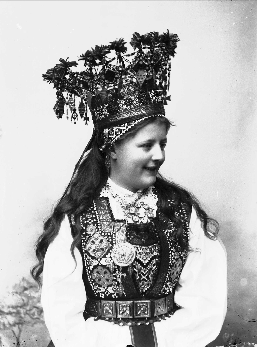 Solveig Lund, Studiofotografi av kvinne med brudedrakt og brudekrone Sittende forann malt bakgrunn med blomster/plante motiv. Hofteutsnitt. Vendt mot høyre. Ser ut av bildet.