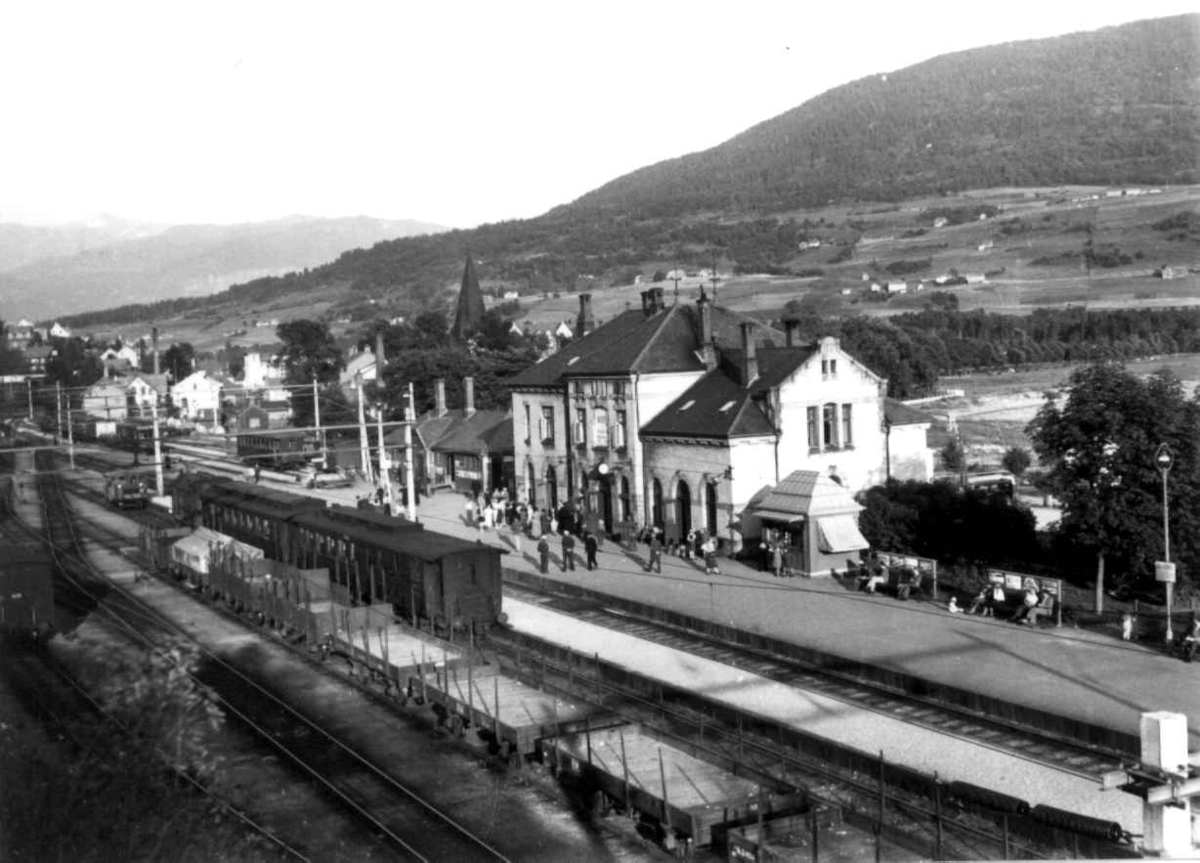 Hermann Christian Neupert, Voss jernbanestasjon 1939. Jernbanevogner og godsvogner på stasjonen. Passajserer på perrongen. Landskap med gårder, jorder og åkrer. Skog og fjell i bakgrunnen.