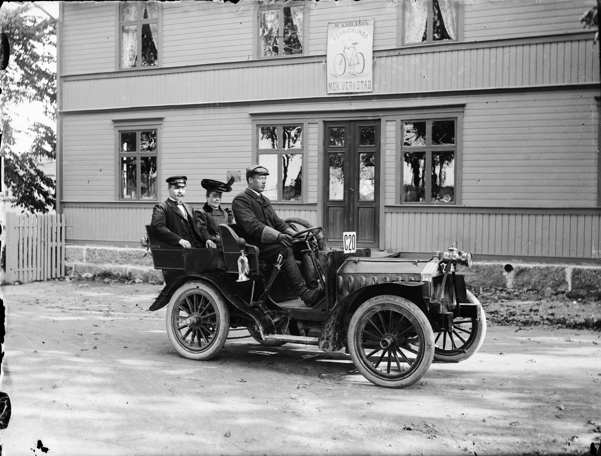Skötsner-Edhlund, Mattias Karlsson i sin bil med en kvinna och chaufför Gustav Lundström utanför "M. Karlsson förnicklings Mekaniska Verkstad", Östhammar, Uppland 1915