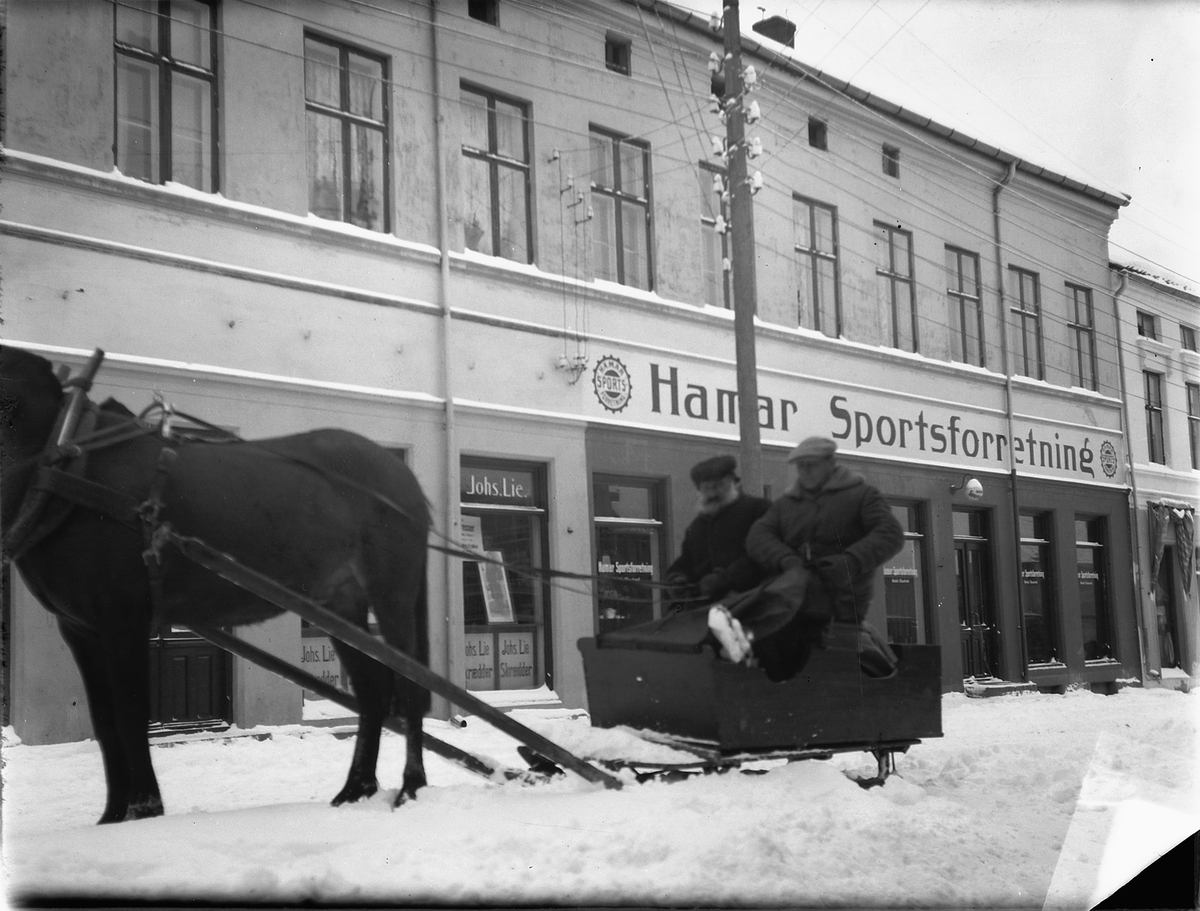 Ragnvald Andersen, Hesten Trygg med Oluf Danielsen og Otto Sinnerud i slede i Torggata i Hamar. Hamar Sportsforretning i bakgrunnen.