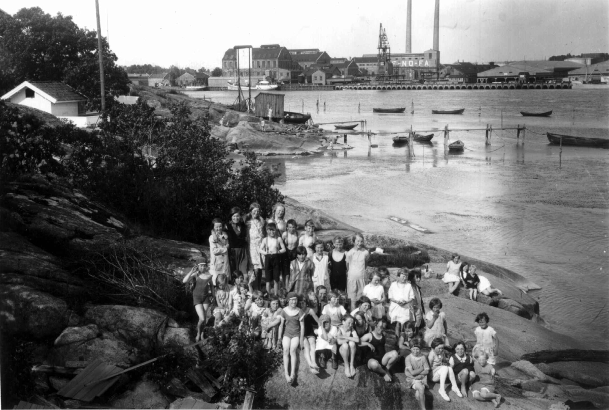 Hermann Christian Neupert, Badestrand på Kråkerøy i Fredrikstad med Denofa Fabrikker i bakgrunnen, antakelig. Barn og voksne i forgrunnen. .