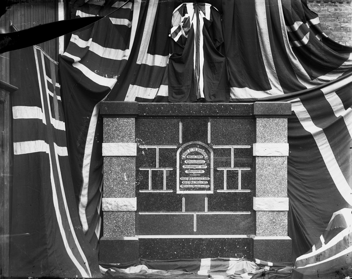 Marthinius Skøien, Gravmonument over John F. A. Køltzow, grunnlegger av "Stenhuggeri med Maskindrift", Akersveien 06-10, Hammersborg, Oslo. Monumentet er oppstilt i bedriftens gård omgitt av norske flagg. Se også NF.32713-015 ved trebygningen til venstre.