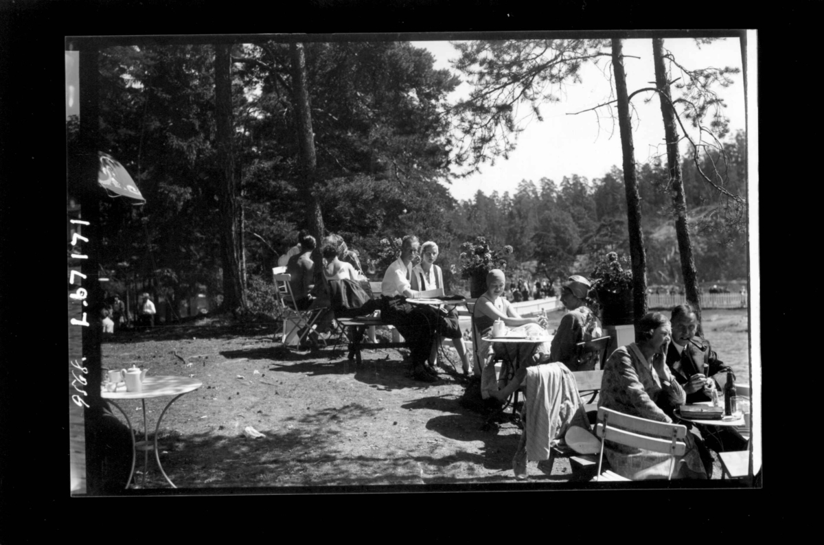 Hermann Christian Neupert, Uteservering. Kvinner og menn ved bordene. Servise på bordene. Skog med stier i bakgrunnen. Parkert bil.