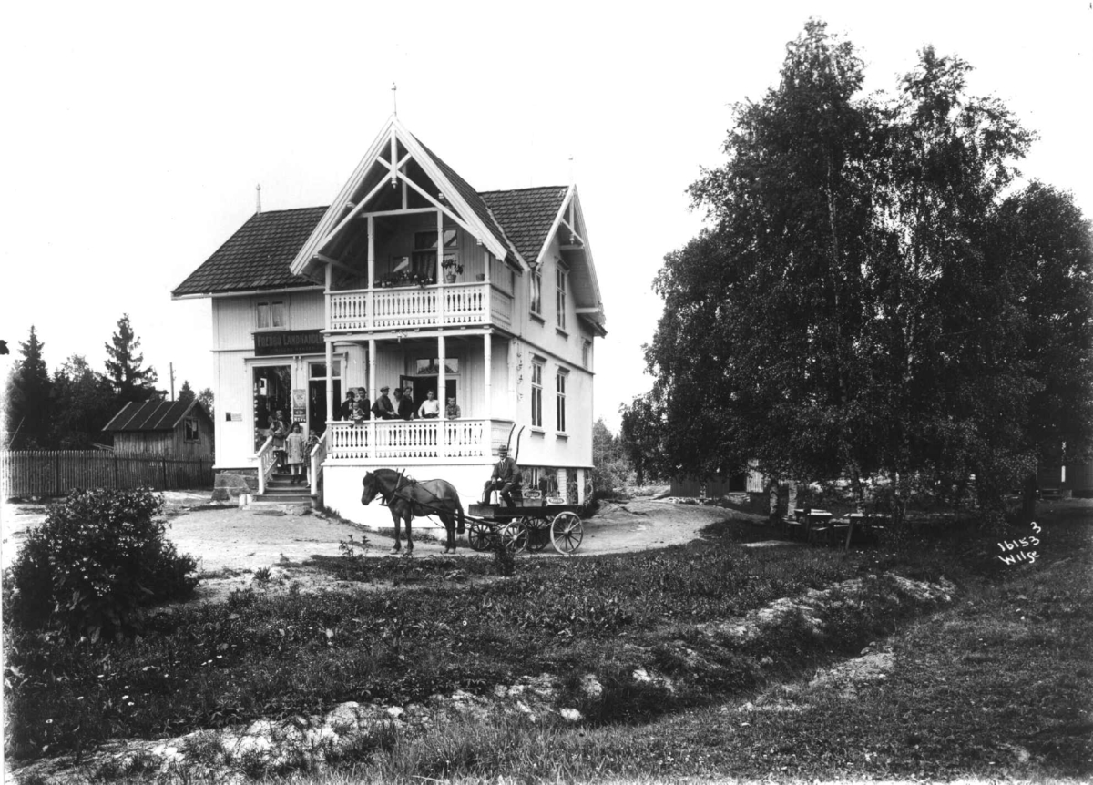 Anders Beer Wilse, Fredbo landhandleri på Høybråten i Oslo. Eksteriør av huset med folk på verandaen og hestekjøretøy utenfor.