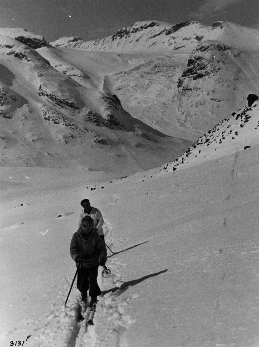 Anders Beer Wilse, Skiløpere ved Galdhøpiggen i Jotunheimen med snødekte fjell og sol.