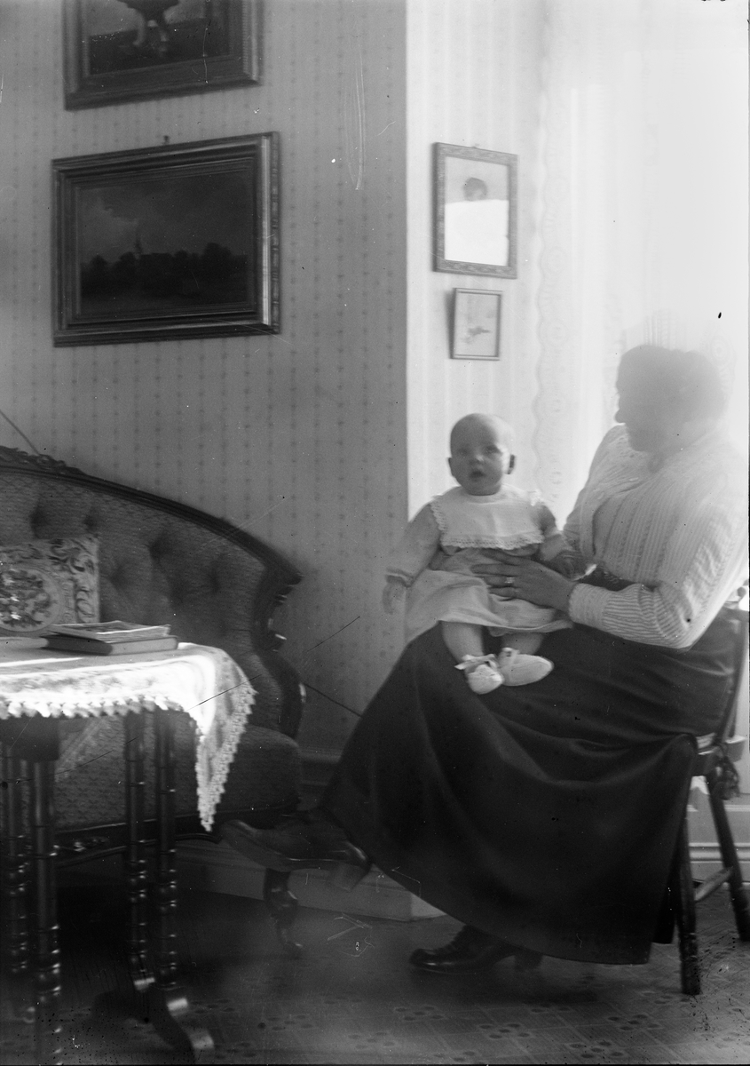 Okänd,, Kvinna sittande framför fönstret med litet barn i famnen i hemmet, sannolikt Sverige sekelskiftet 1900