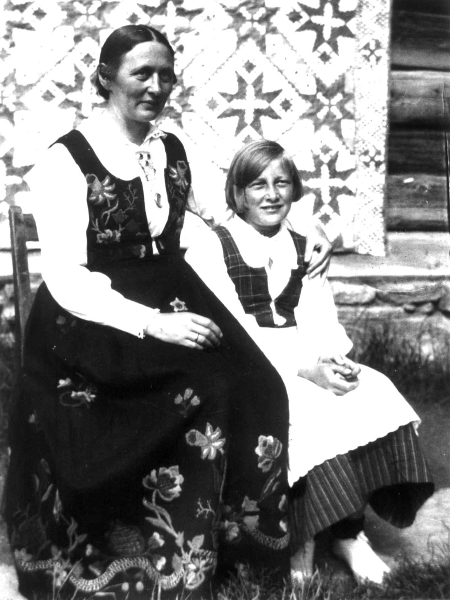 Anna Grostøl, Antagelig Lesja, Oppland 1934. To ukjente kvinner kledd i bunader sittter foran en husvegg med et teppe på.