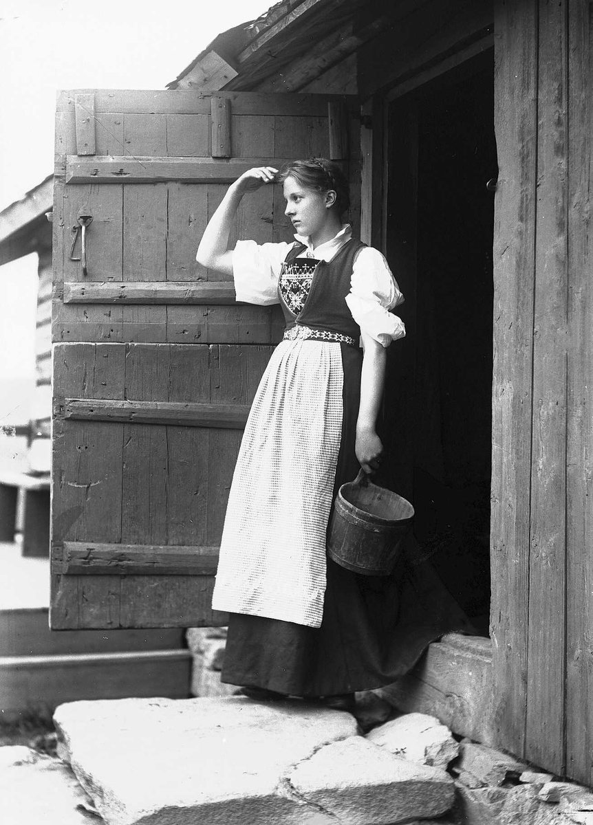 Solveig Lund, Kvinne i Hardangerdrakt står i inngangsdøren til seter. Holder en bøtte i hånden.