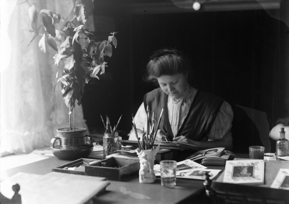 Solveig Lund, Antakelig Solveig Lund selv i studioet sitt arbeidene med kolorering av papirfotografier. Fotografier og pensler på pulten.