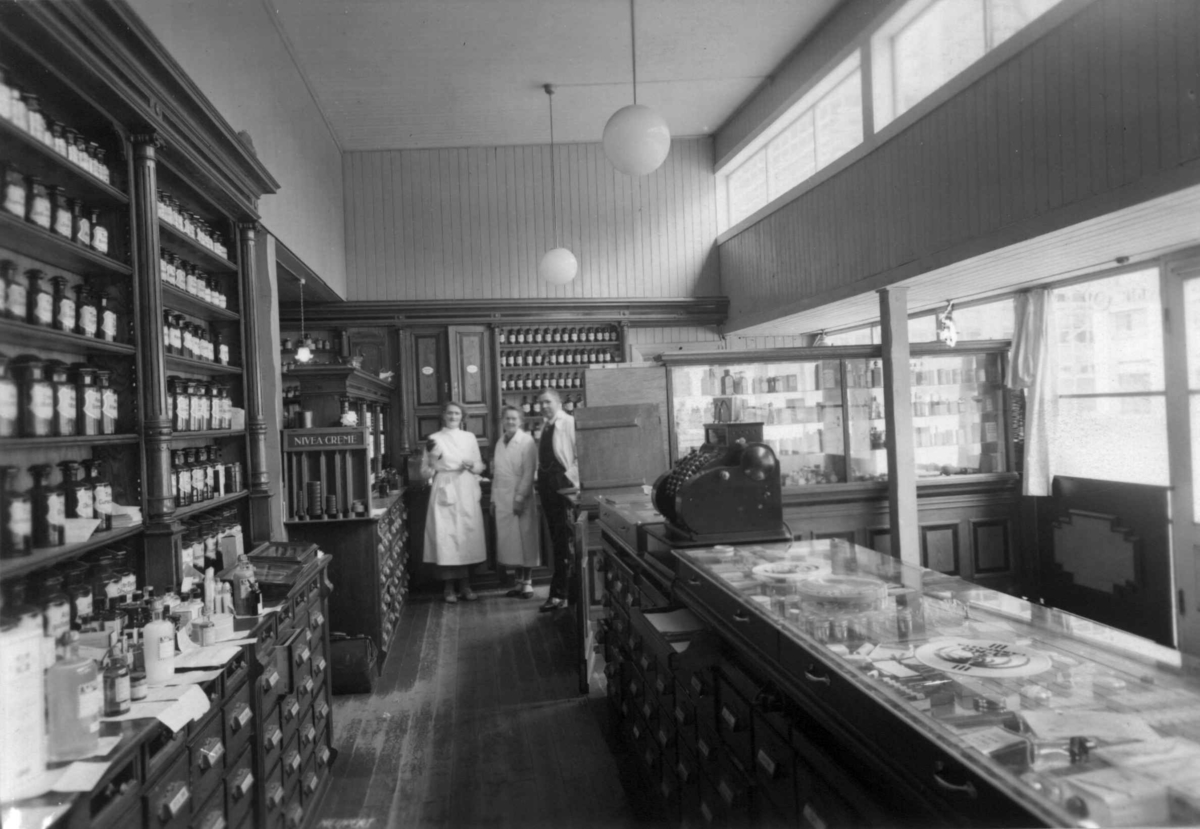 Hermann Christian Neupert, Rådhusapoteket, Oslo 1935-1937. Interiør. Disk med skuffer. Kassaapparat. Hyller med apotekervarer på flasker. To kvinner og en mann i bakgrunnen.