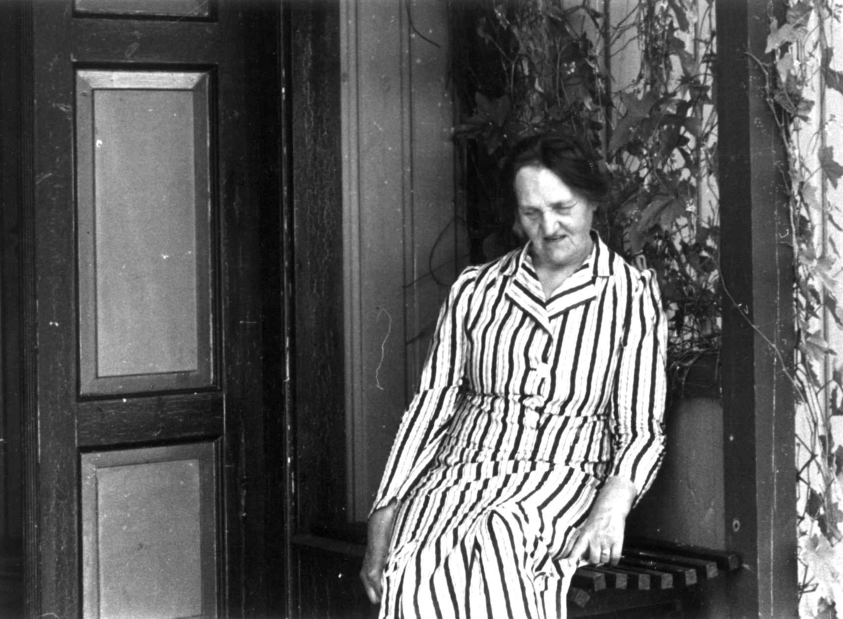 Anna Grostøl, Dølemo Åmli, Aust-Agder 1940. En kvinne i stripete kjole sitter på en benk ved døren til et inngangsparti.