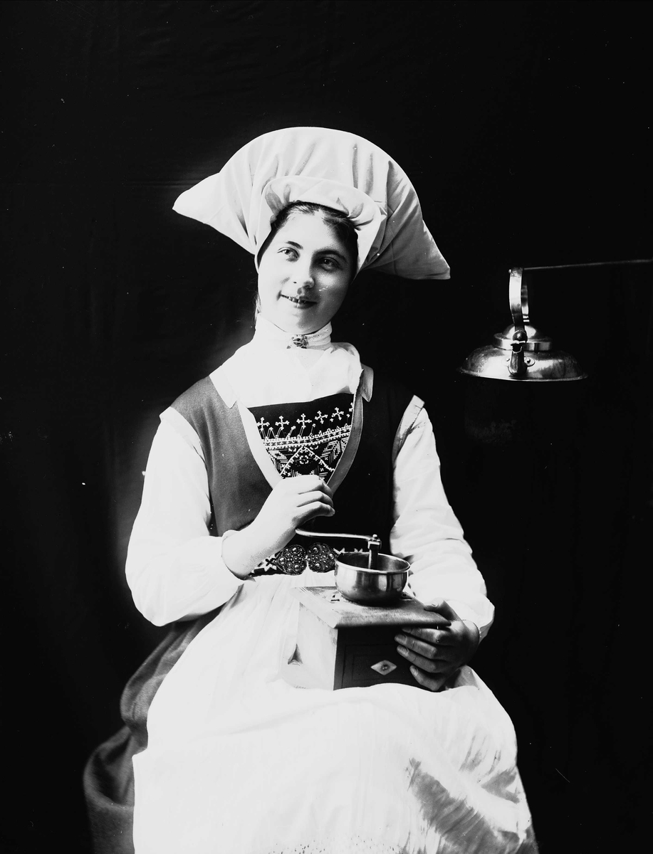 Solveig Lund, 1/2 portrett av kvinne i drakt og skaut, tatt i fotografens studio. Hun sitter og holder en kaffekvern på fanget. Til venstre for henne henger en kaffekjele. Nøytral bakgrunn.