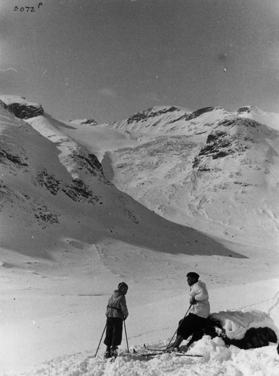 Anders Beer Wilse, Skiløpere ved Galdhøpiggen i Jotunheimen med snødekte fjell og sol.