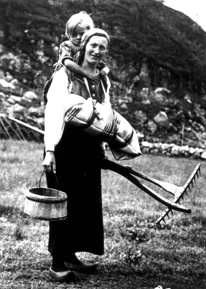 Anna Grostøl, Oline Seldal med barn i fatle, i Sirdal kalt linde, et barn på ryggen og bøtte og rive i hendene, Fidjeland, Sirdal, Vest-Agder 1936. Rekonstruksjon. Barnet hun har på ryggen er hennes niese, Tordis Harbo, f. Fidjeland (f. 1932).Et hus i bagrunnen med kvinne og barn i inngangspartiet. Oline Seldal …