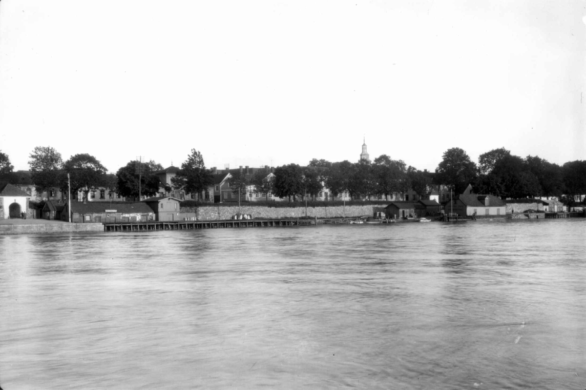 Hermann Christian Neupert, Fredrikstad. 1930. Bebyggelse sett fra elva. Trær langs elvekantern. Fortøyde båter langsmed kaia.