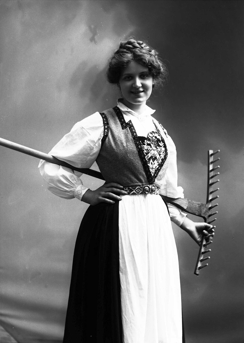 Solveig Lund, 3/4 dels studioportrett av kvinne i drakt, med oppsatt hår. Hun holder en rive bak ryggen. Bakgrunnen er nøytral.