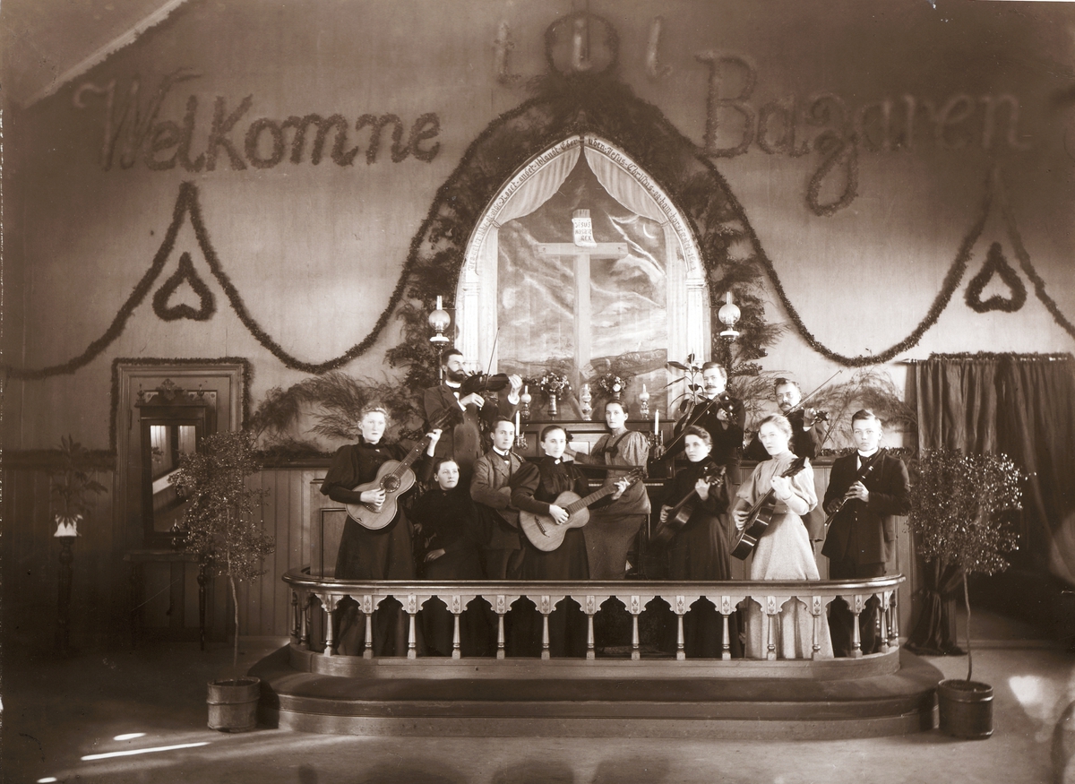 Ukjent, Metodistkirken Hamar, Interiør. Strengemusikken i Metodistkirken Hamar omkring 1895. Alteret, alteringen pyntet. ”Welkomne til Bazaren”. Personene på bildet er fra venstre : Emma Larsen Røtvold, Gina Jensen, pastor Johs. Lie (1894-1897), Gjermund Johnsen, Marie Johnsen, ukjent, Gustava Olsen, kjøpm…