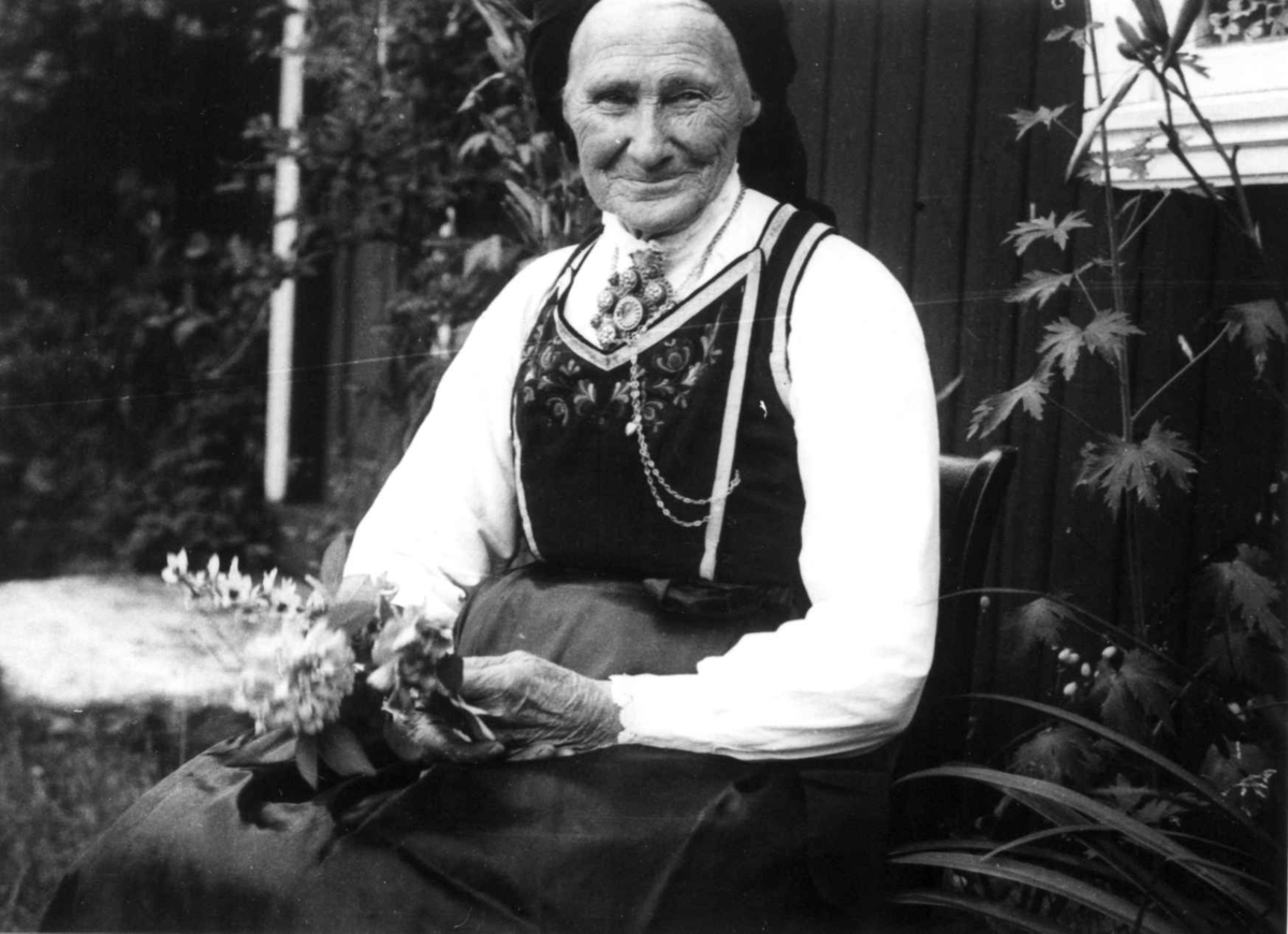 Anna Grostøl, 1935. Portrett av en kvinne som sitter med blomster på fanget utenfor en husvegg.