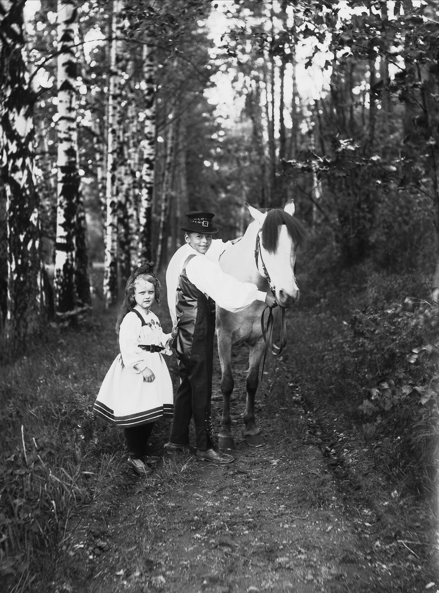 Solveig Lund, Gutt og pike i nasjonaldrakt som står på skogsti. Gutt holder en hest i tømmene, piken holder gutten i vesten.