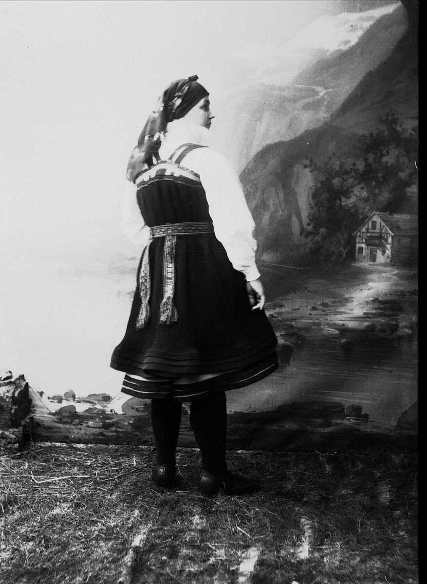 Solveig Lund, Studioportrett av kvinne i nasjonaldrakt avbildet bakfra. Står på gress/jordbunn Malt bakgrunn med fjord og fjell og et lite hus.