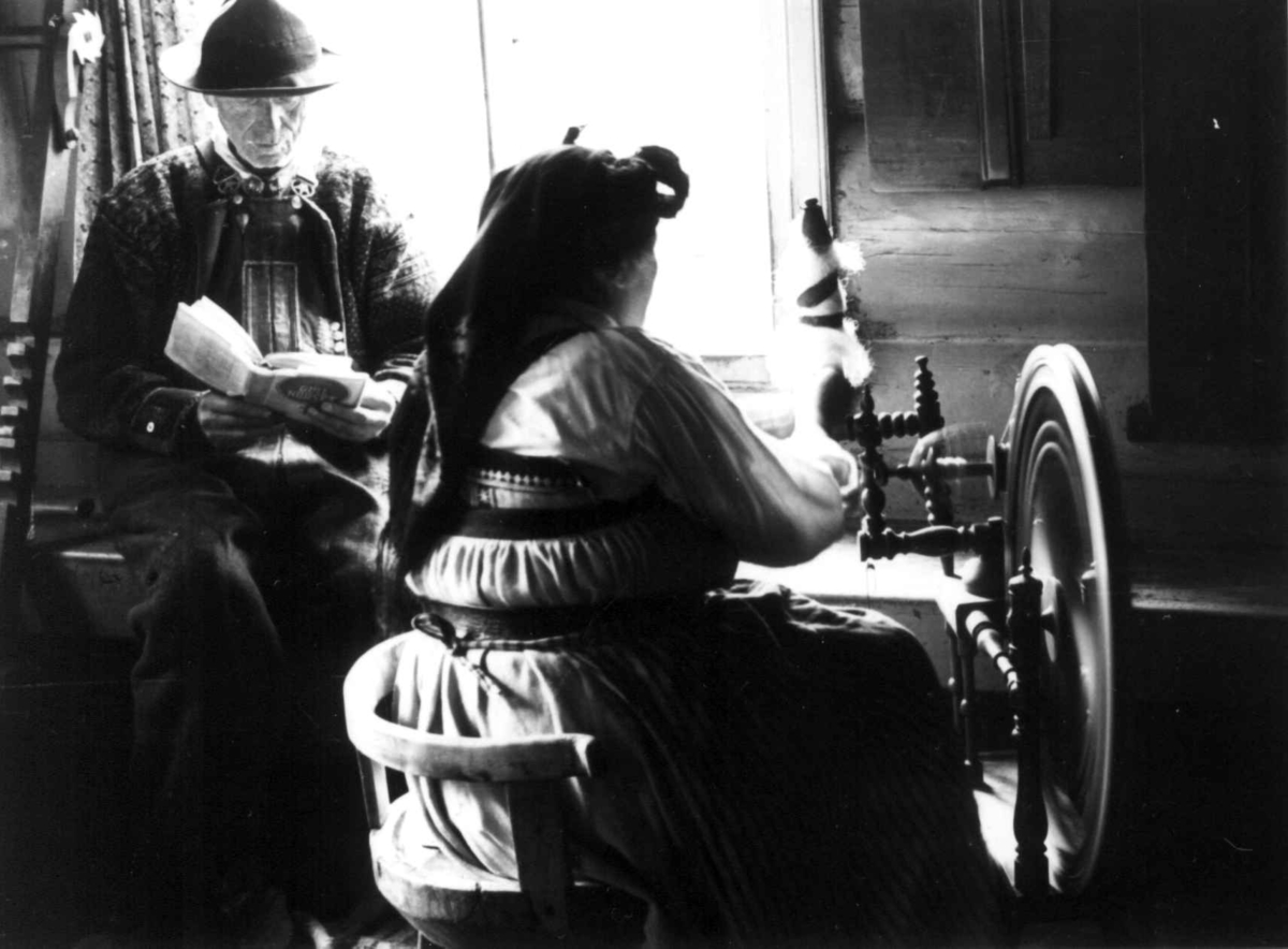 Anna Grostøl, Valle, Aust-Agder 1938. Spinning. Gunnhild sitter ved rokken og Typpe-spinner til varp. hennes mann Håvar sitter ved siden av og leser i en bok.