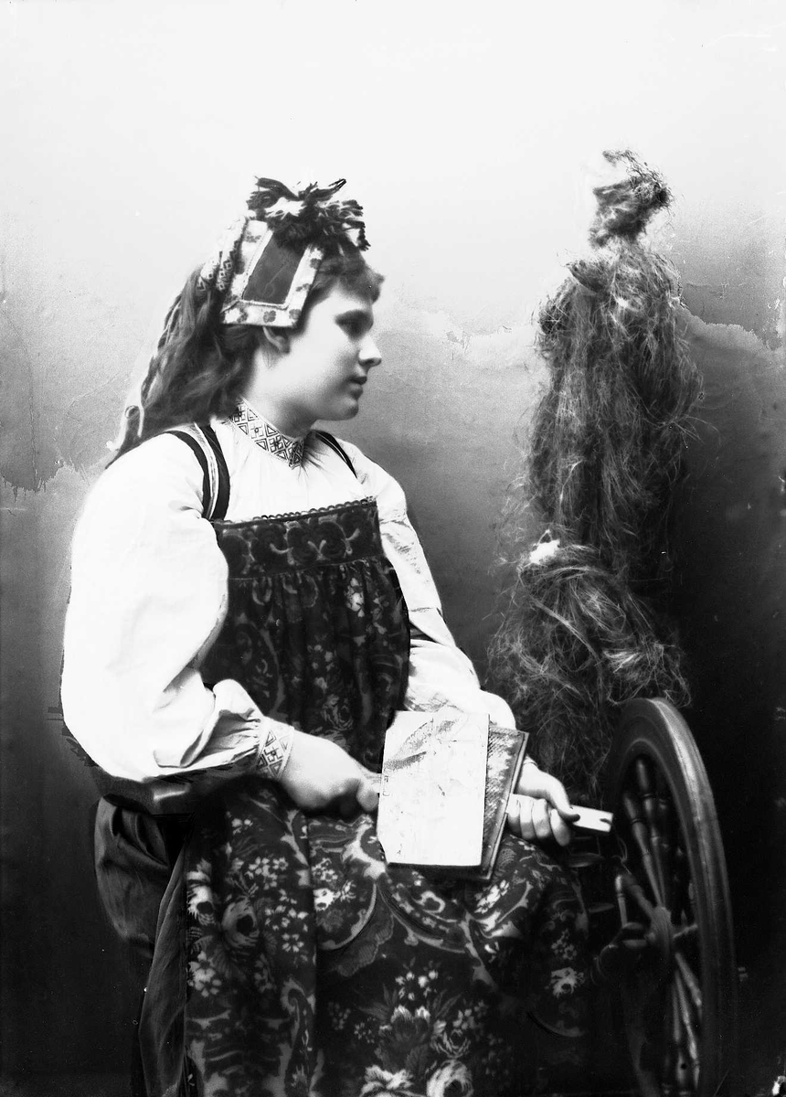Solveig Lund, Studioportrett av kvinne i drakt og skaut, som sitter ved en rokk og karder. Bakgrunnen er malt.
