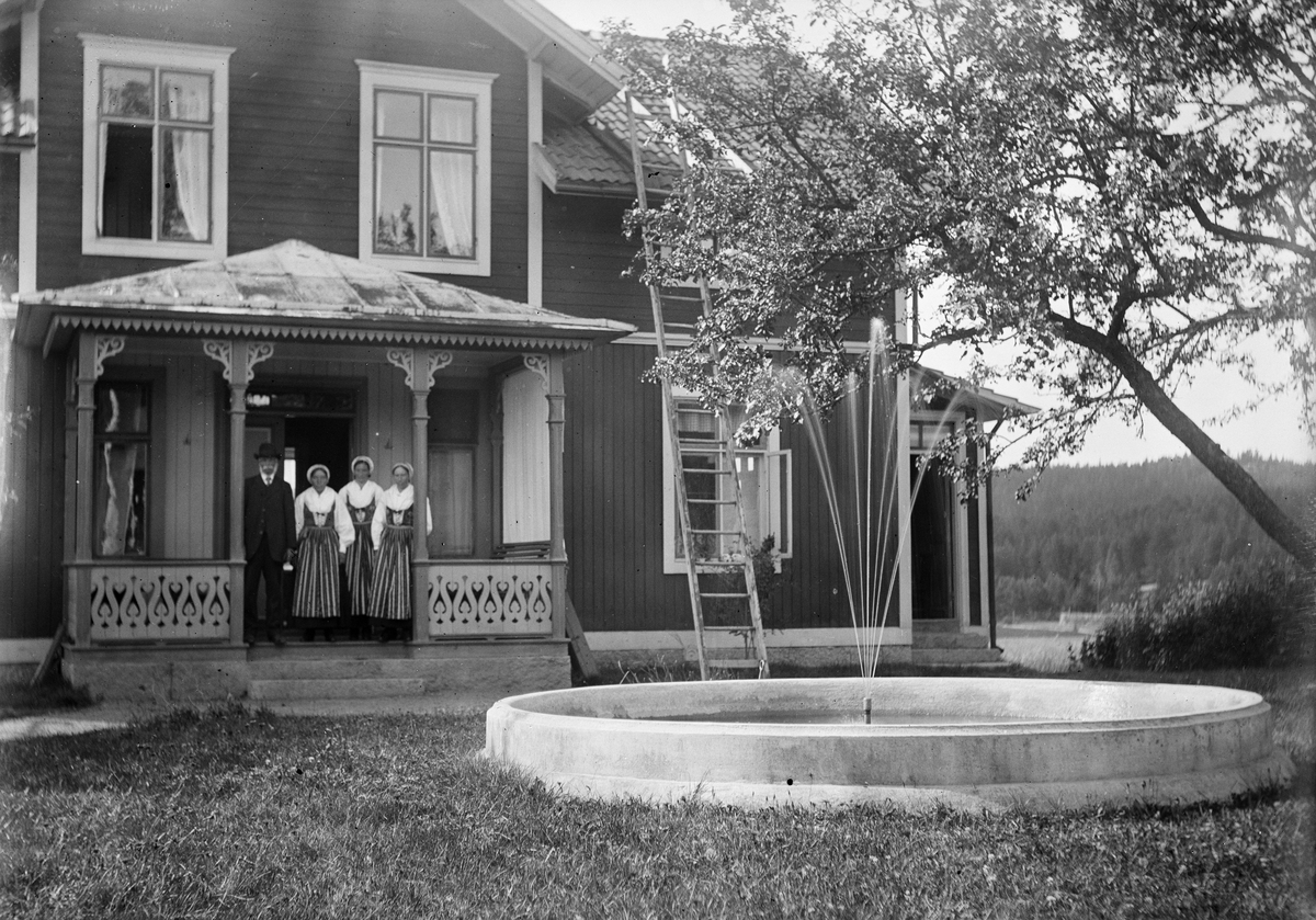 Okänd,, Svens Olle with the daughters Anna, Britta and Kristina, dressed in folk costume, standing in porch to residential buildings, in front of the house there is a fountain, Svensgården, Dalarna, Midsummer 1919