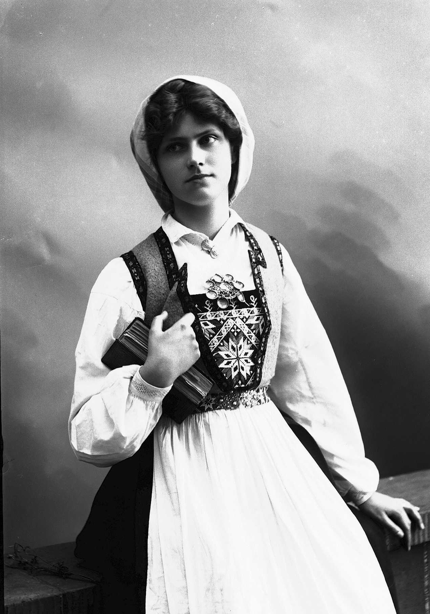 Solveig Lund, Studioportrett av kvinne i drakt og skaut. Hun sitter på en benk og holder en bok i høyre arm. Bakgrunnen er nøytral.